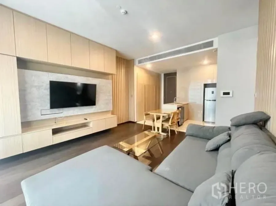 Condominium for rent in Watthana, Bangkok - Modern living room with grey sectional sofa, built-in TV wall and open kitchen.