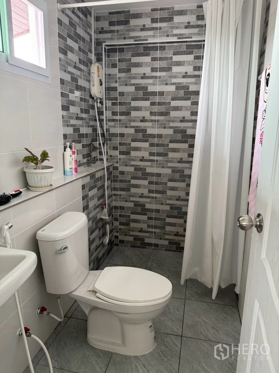 Detached House for rent in Saphan Sung, Bangkok - Compact bathroom with patterned gray tiles, window, toilet, sink, water heater, and shower curtain.