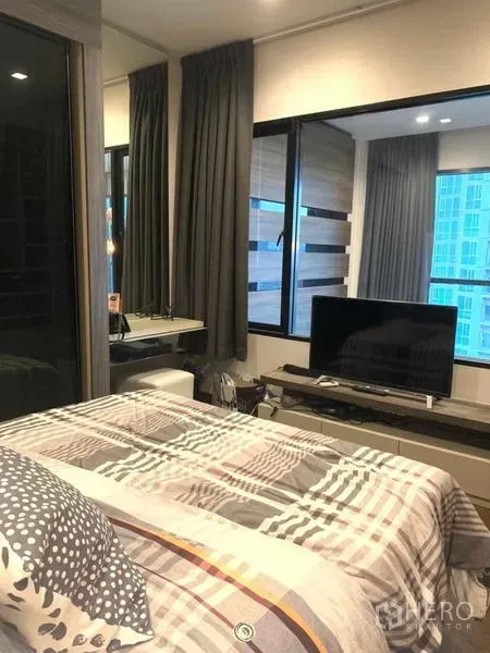 Condominium for rent or sale in Huai Khwang, Bangkok - Cozy bedroom with large windows, TV console, and built-in vanity overlooking city towers.