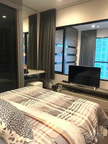 Condominium for rent or sale in Huai Khwang, Bangkok - Cozy bedroom with large windows, TV console, and built-in vanity overlooking city towers.