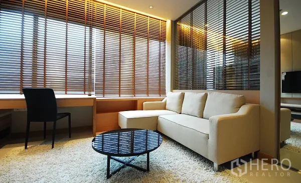 Condominium for rent in Khlong Toei, Bangkok - Bright condo living room with cream sofa, glass coffee table, shag rug and wide wooden blinds beside a built-in work desk.