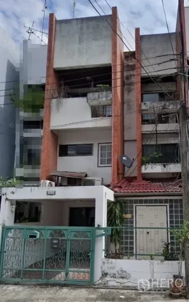 店屋 出售在Khlong Toei, Bangkok - 曼谷临街多层商铺式联排外观，红砖立柱与混凝土立面，带绿色金属大门和车位，阳台有绿植，可见街边电线与相邻建筑。