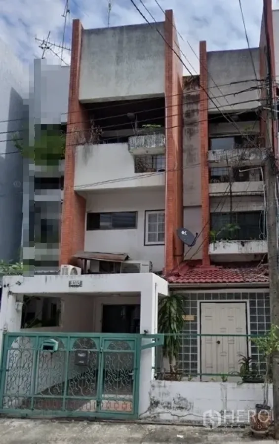 店屋 出售在Khlong Toei, Bangkok - 曼谷临街多层商铺式联排外观，红砖立柱与混凝土立面，带绿色金属大门和车位，阳台有绿植，可见街边电线与相邻建筑。