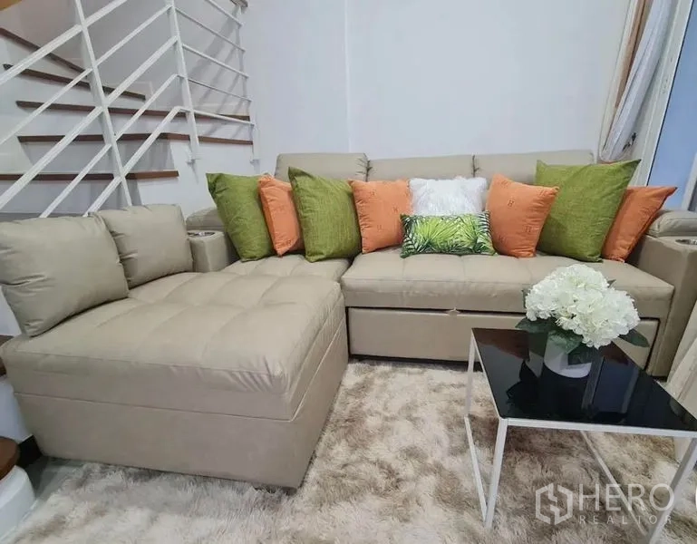 联排别墅 出租在Prawet, Bangkok - 舒适客厅，米色转角沙发、缤纷抱枕、长绒地毯与楼梯。