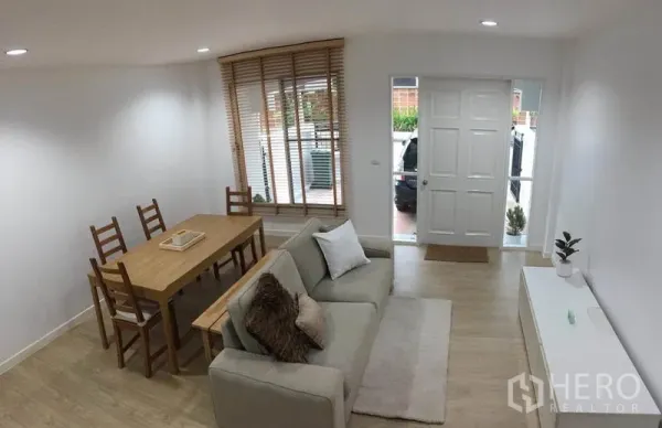 ทาวน์เฮาส์ ให้เช่าในLat Phrao, Bangkok - มุมมองห้องรับแขกไปยังประตูทางเข้า มีชุดโซฟา ชุดโต๊ะกินข้าว และมู่ลี่หน้าต่าง