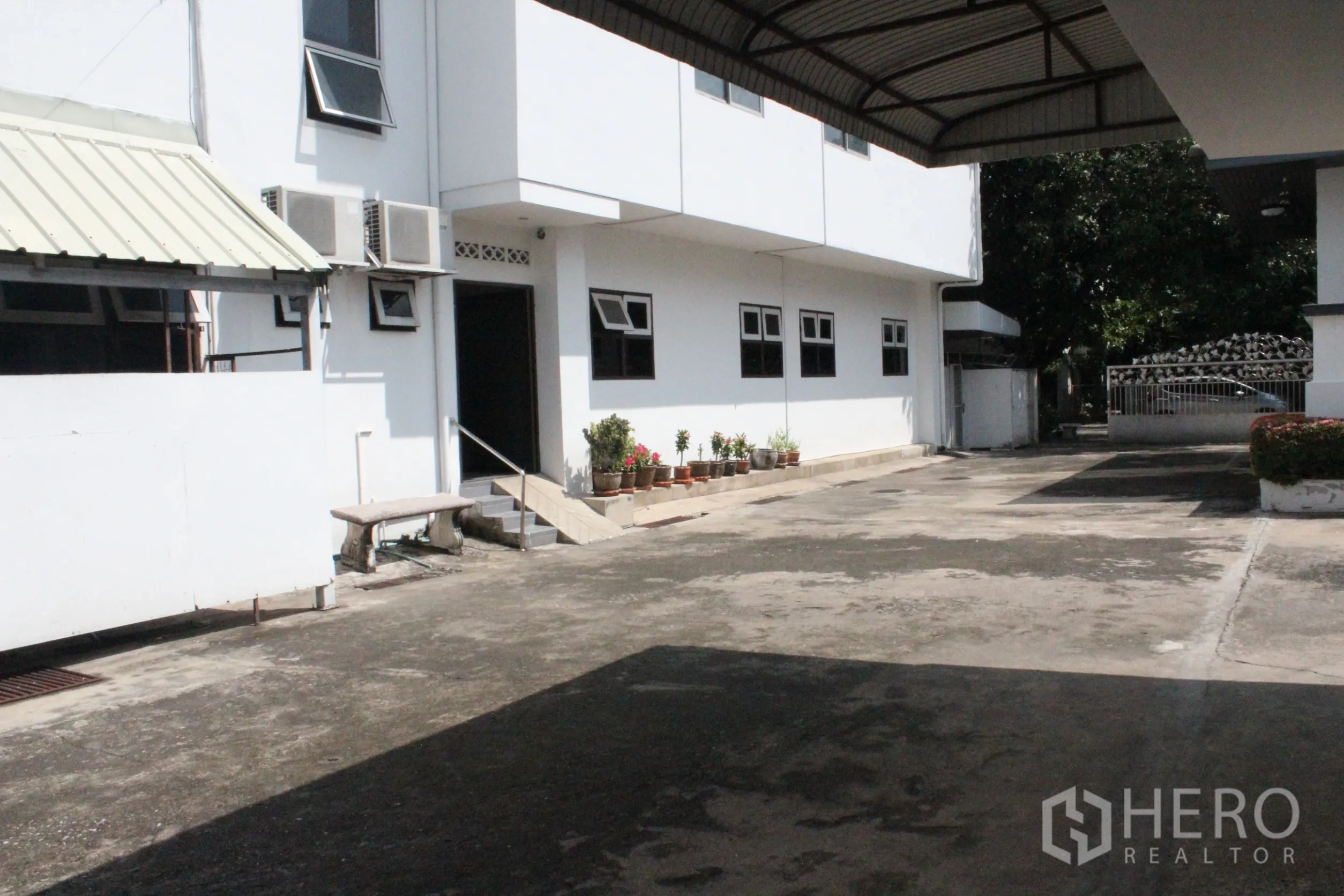 Home Office for rent in Khlong Toei, Bangkok - Spacious concrete driveway and white low-rise building with covered carport, potted plants and external air-conditioning units.