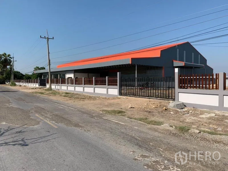 Warehouse for sale in Phra Nakhon Si Ayutthaya - Street view of a modern gray-and-orange warehouse with a secure perimeter wall and power lines in rural Ayutthaya.