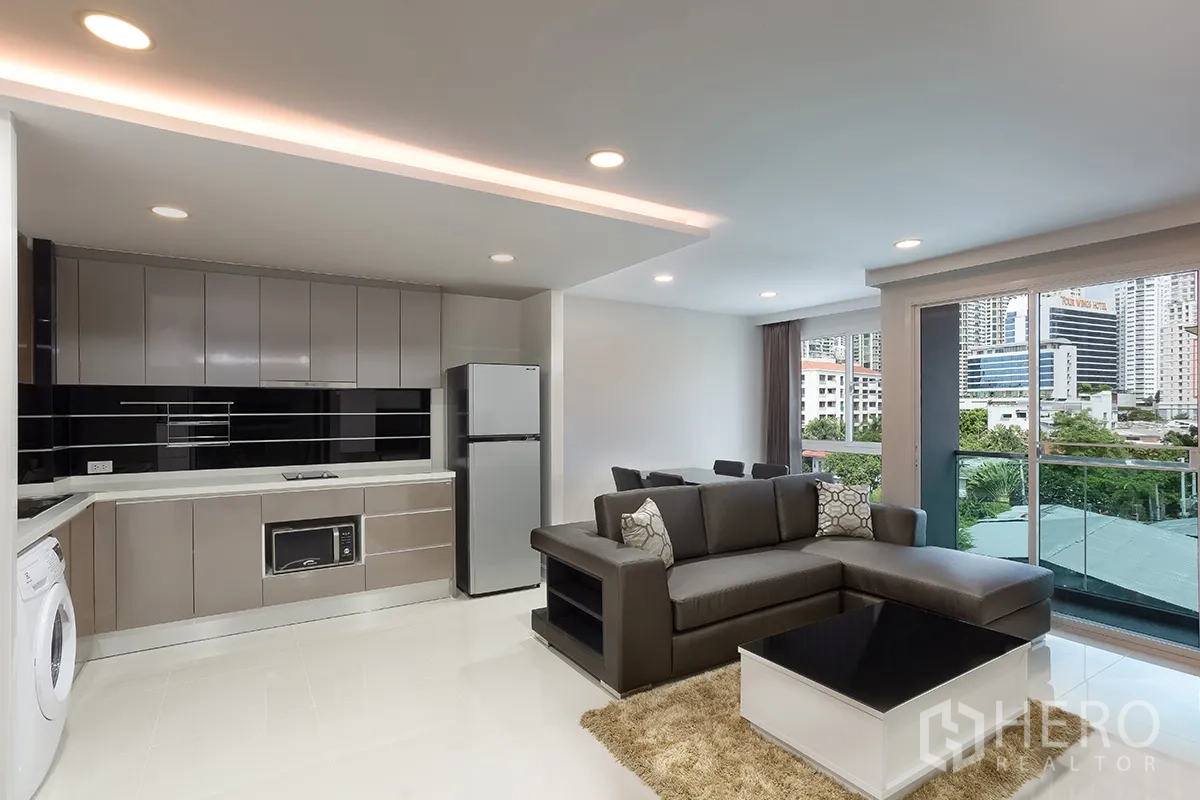 Condominium for rent in Khlong Toei, Bangkok - Open-plan living room with L-shaped sofa, dining area, balcony access, and fitted kitchen.