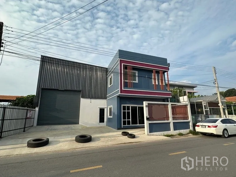 货栈 出租在Lam Luk Ka, Pathum Thani - 正立面视角：金属板仓库及现代蓝色办公楼，带车道与路边出入口