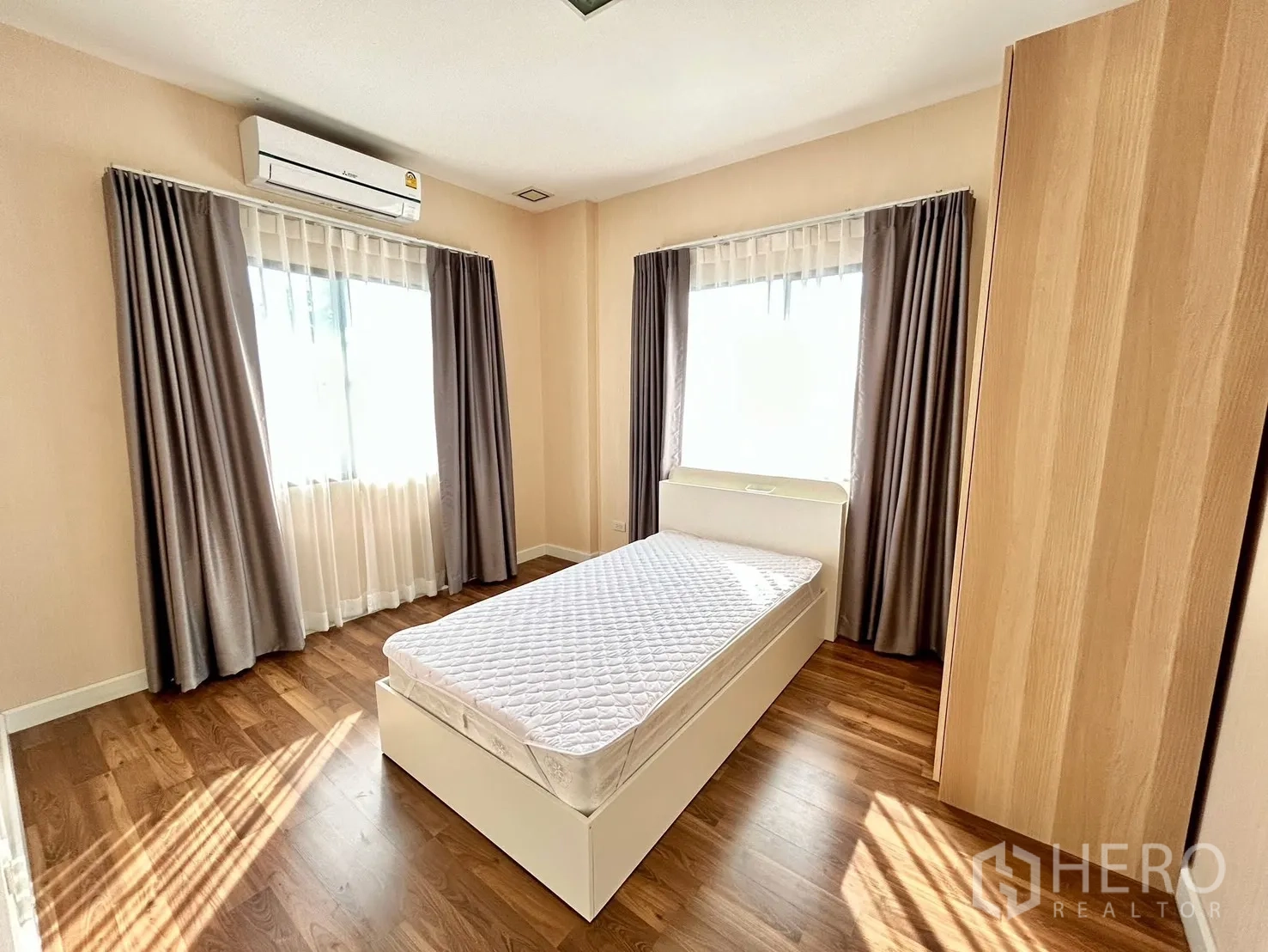 Detached House for rent or sale in Prawet, Bangkok - Bright bedroom with a single bed, wooden flooring, two curtained windows and a wall-mounted air conditioner.