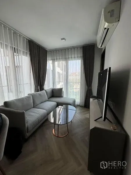 Condominium for rent in Watthana, Bangkok - Living room with sofa, TV console and floor-to-ceiling windows opening to a balcony.