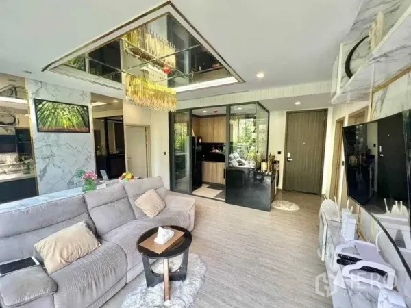 Condominium for sale in Watthana, Bangkok - Spacious lounge with gray sofa, glass-walled kitchen, marble TV wall and statement chandelier.