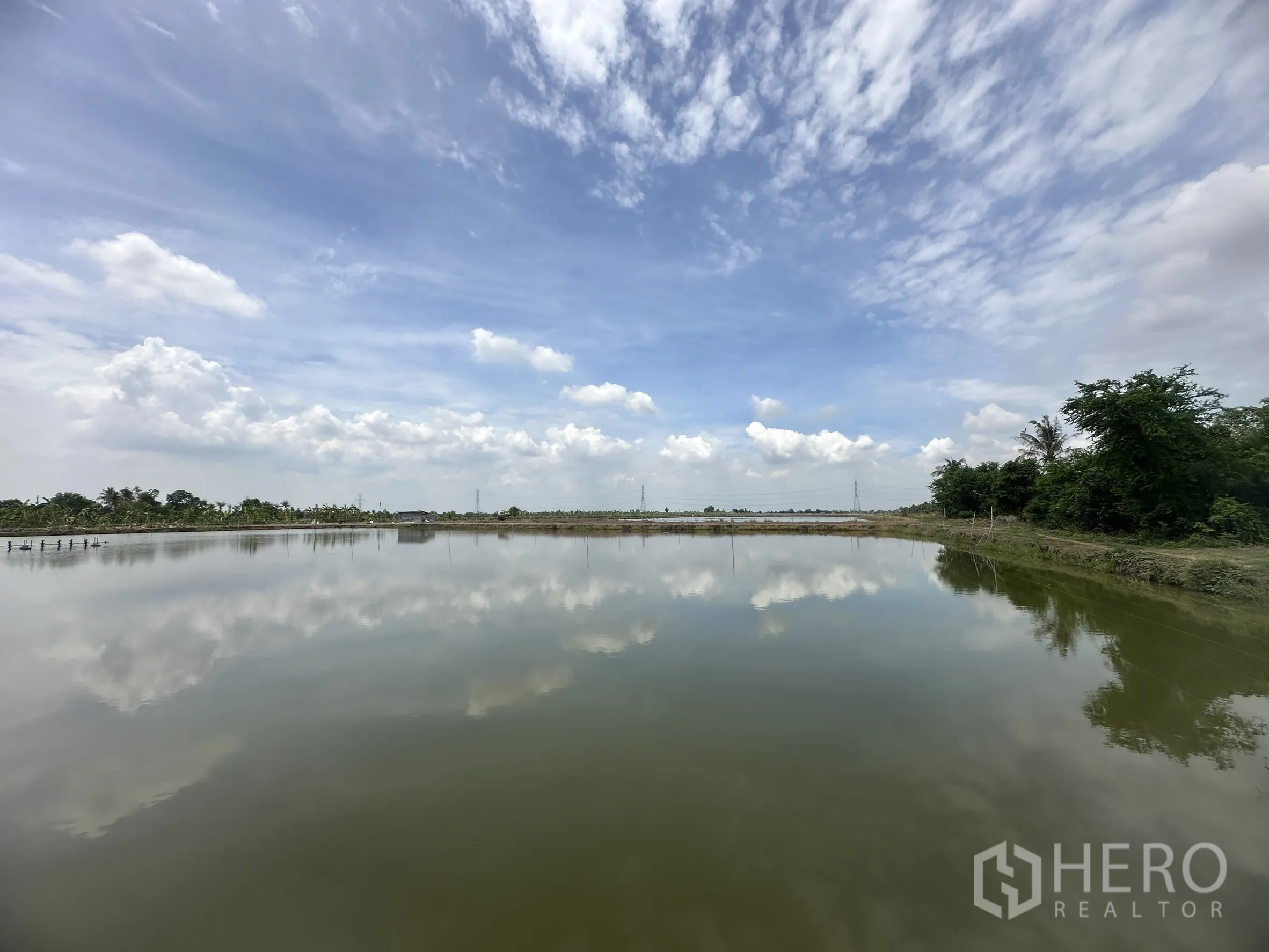 Land for sale in Bang Nam Priao, Chachoengsao - Large water reservoir bordered by greenery and levee paths under a cloudy sky.