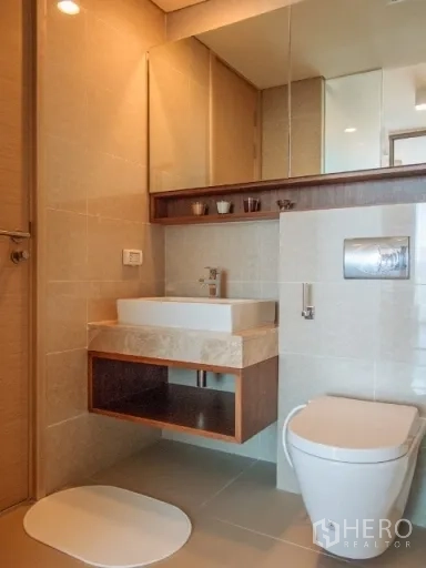 Condominium for rent in Watthana, Bangkok - Modern bathroom with wall-mounted vanity, large mirror cabinet, and toilet in neutral tiles.
