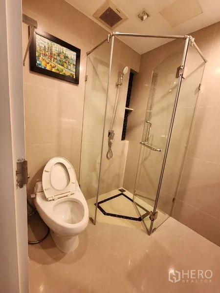 公寓 出租在Bang Rak, Bangkok - 米色浴室，玻璃淋浴间、手持花洒与坐便器，墙上装饰画。