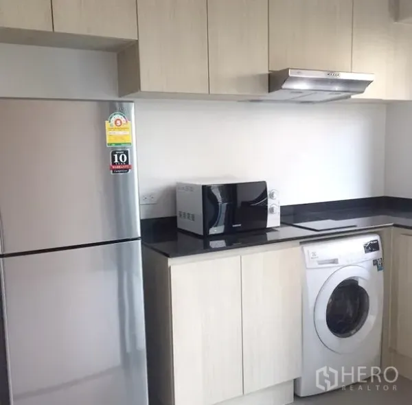 Townhouse for rent in Suan Luang, Bangkok - Fitted kitchen with light wood cabinets, black countertop, stainless fridge, microwave and front‑load washer.