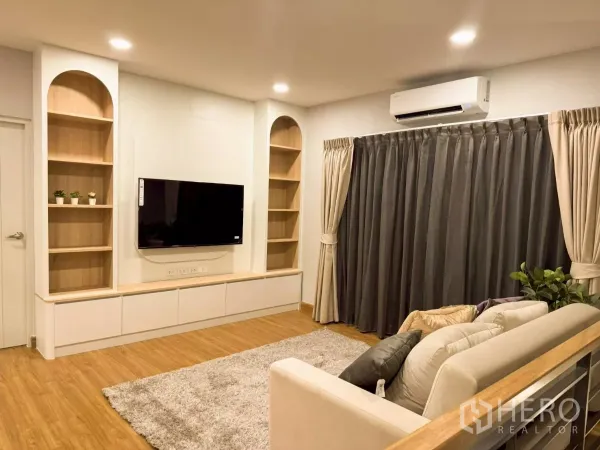 独立式住宅 出租在Bang Phli, Samut Prakan - 客厅设壁挂电视与嵌入式书架，木地板与遮光窗帘，灯光温暖。