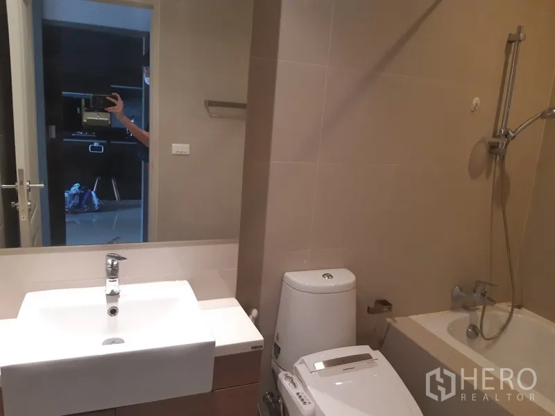 Condominium for rent in Bangkok - Bathroom with bathtub and handheld shower next to vanity and toilet.