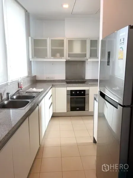 公寓 出租在Khlong Toei, Bangkok - 明亮的走廊式封闭厨房，白色柜体、花岗岩台面、嵌入式烤箱和大冰箱。