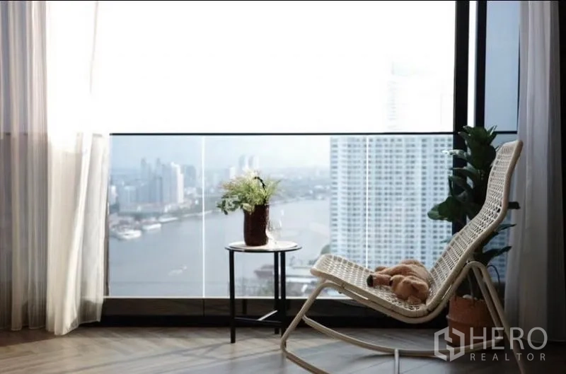 คอนโด ให้เช่า - มุมนั่งเล่นริมกระจกพร้อมเก้าอี้หวายและโต๊ะข้างหันสู่แม่น้ำ