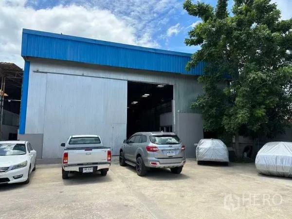 货栈 出租在Mueang Pathum Thani, Pathum Thani - 仓库立面，宽大卷门、现场停车位与遮阴大树
