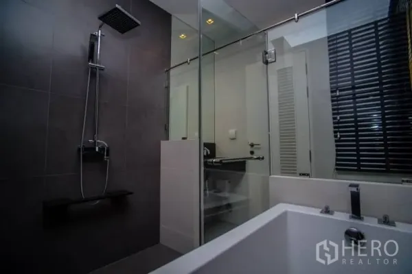Condominium for rent in Khlong San, Bangkok - Contemporary bathroom featuring a glass shower with rain head and a separate bathtub with dark tile walls.