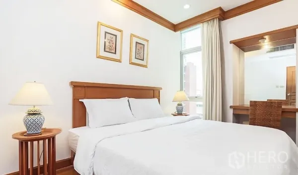 Condominium for rent in Pathum Wan, Bangkok - Cozy bedroom with white bedding, wooden headboard, bedside lamps, window, and a built-in vanity.