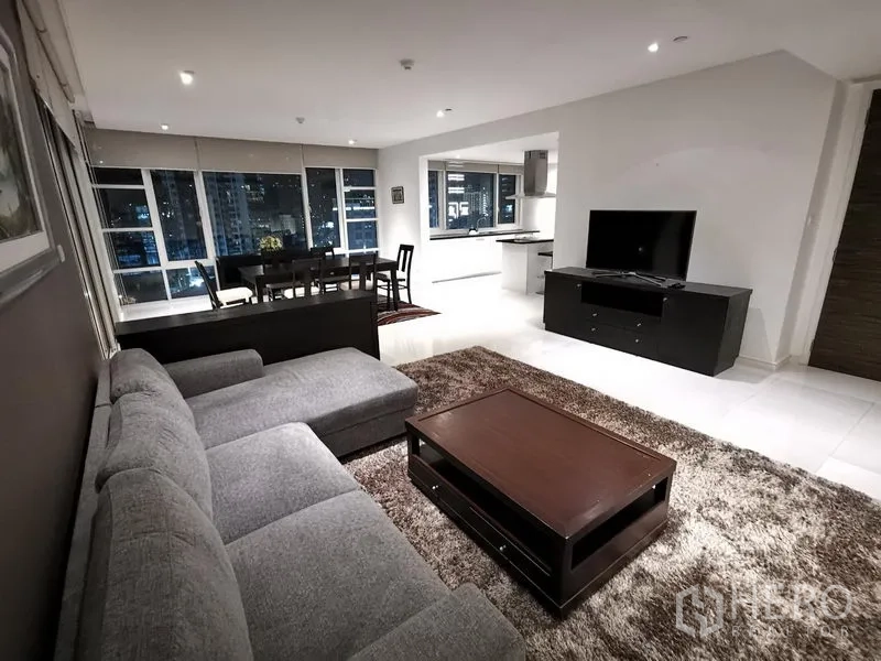 Condominium for rent in Watthana, Bangkok - Living area facing the dining space and open kitchen with floor-to-ceiling windows at night.