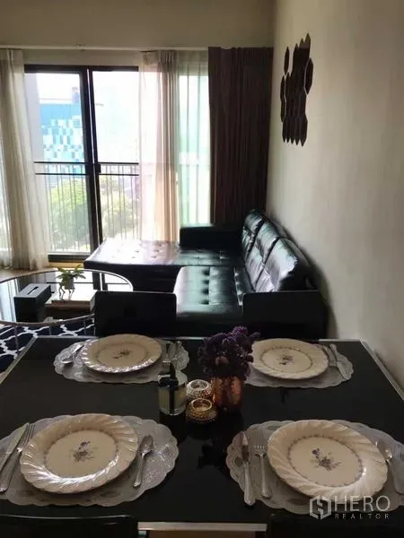 Condominium for rent in Watthana, Bangkok - Bright living and dining area with a table set for four, black leather sofa, and balcony with sheer curtains.