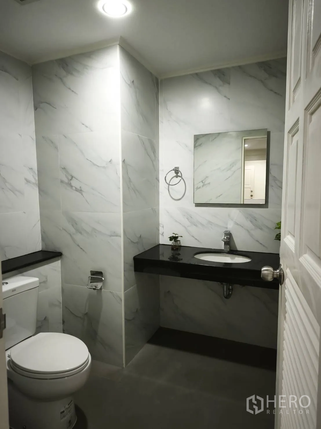 Condominium for rent in Watthana, Bangkok - Bathroom with marble-effect wall tiles, black countertop vanity, toilet, and mirror.