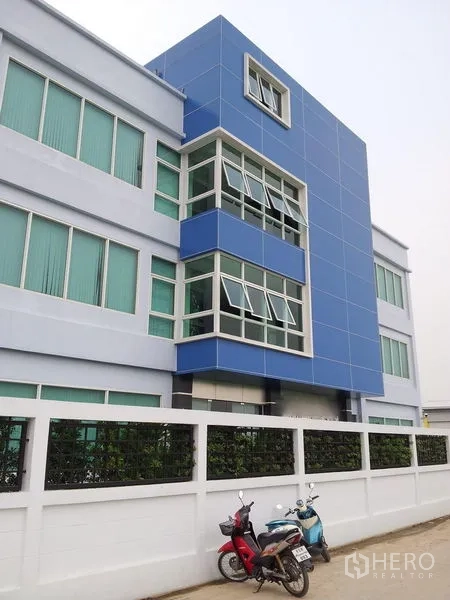货栈 出租或出售在Bang Phli, Samut Prakan - 蓝白配色四层仓库与办公楼外立面，转角大窗，前有白色围墙。