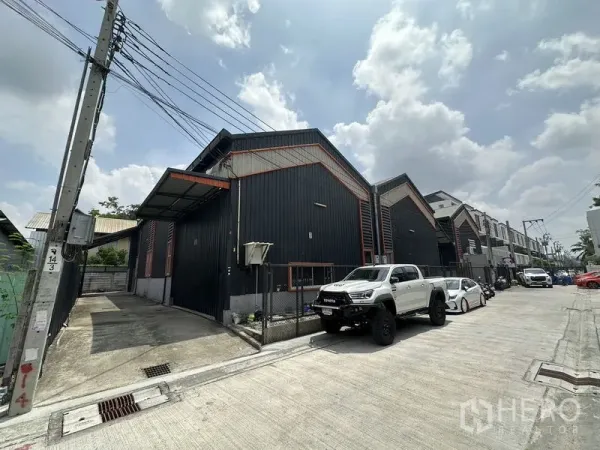 คลังสินค้า ให้เช่าในLat Phrao, Bangkok - ภาพถนนหน้าโกดังผนังเมทัลชีทสีดำ หลังคาจั่ว พร้อมที่จอดรถในซอยเงียบสงบย่านลาดพร้าว