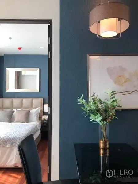 Condominium for rent or sale in Ratchathewi, Bangkok - View from dining nook to a bedroom with blue accent wall, pendant light, framed art, and a vase on the table.