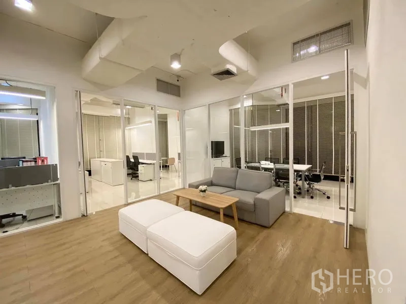 Office Space for rent in Bangkok - Breakout space with sofa and ottoman, surrounded by glass-partitioned rooms and workstations.