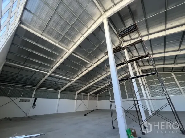 Warehouse for rent in Min Buri, Bangkok - High-ceiling warehouse showing steel rafters and mobile scaffolding for maintenance.