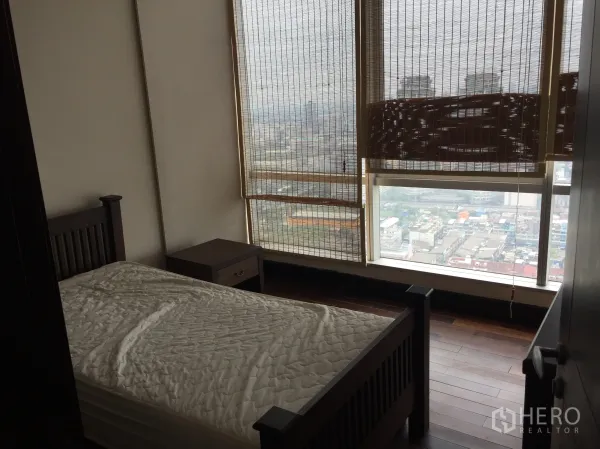 Condominium for rent in Sathon, Bangkok - Bedroom with single bed, wood floors, and large windows with bamboo blinds overlooking the Sathorn skyline.