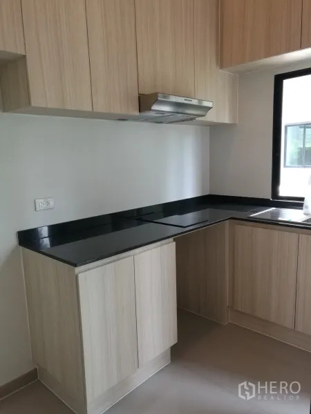 Townhouse for rent or sale in Suan Luang, Bangkok - L-shaped kitchen with light wood cabinets, black countertop and window.