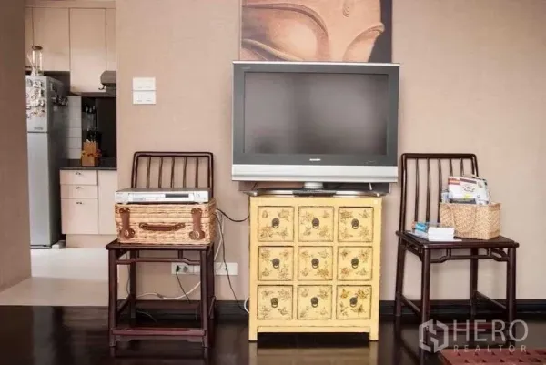 Condominium for rent in Watthana, Bangkok - Living area wall with flat-screen TV on a vintage yellow cabinet, two wooden chairs and kitchen view.