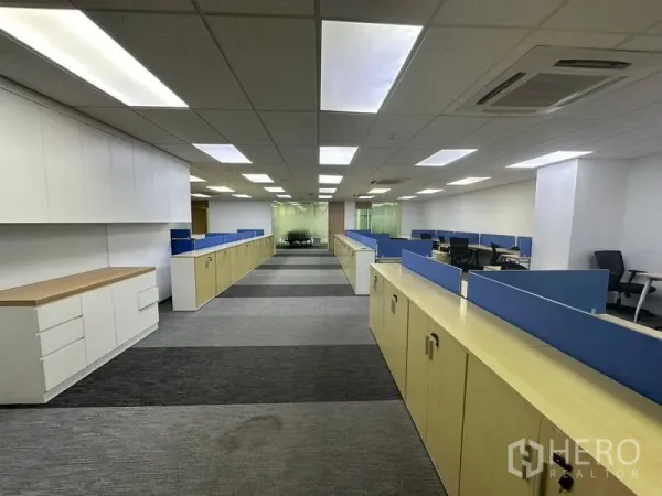Office Space for rent - Central aisle view of twin rows of desks with overhead white cabinetry and a glass-walled meeting room ahead.