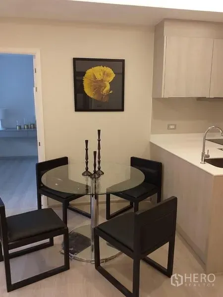 公寓 出租在Khlong Toei, Bangkok - 玻璃餐桌配四把黑色椅子，旁有浅木色一字型厨房与黄色装饰画。