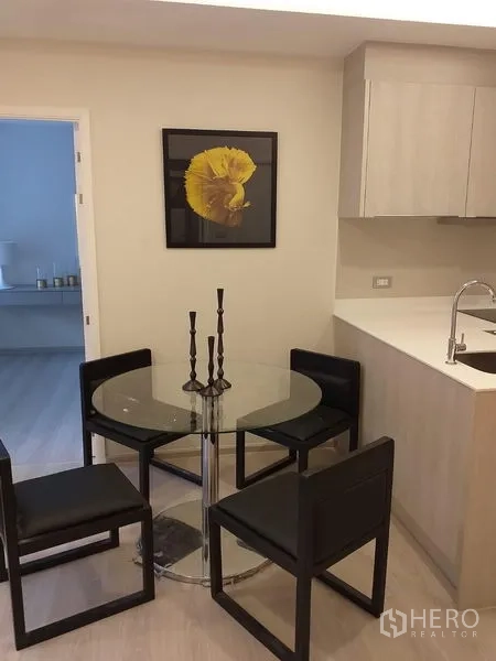 公寓 出租在Khlong Toei, Bangkok - 玻璃餐桌配四把黑色椅子，旁有浅木色一字型厨房与黄色装饰画。