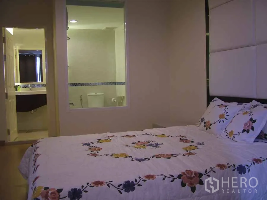 Condominium for rent or sale in Watthana, Bangkok - Bedroom with floral bedspread looking toward an ensuite bathroom through an interior window.