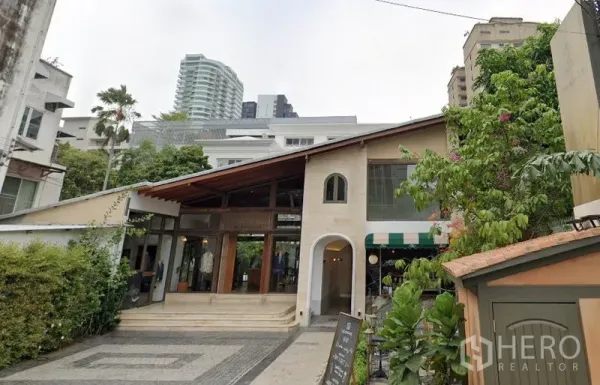 零售空间 出租在Khlong Toei, Bangkok - 两层零售建筑，斜坡屋顶、拱形入口与落地玻璃，前方铺装前庭与绿化，背景为城市高楼。