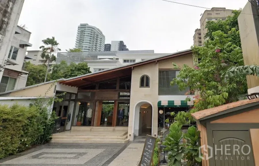 零售空间 出租在Khlong Toei, Bangkok - 两层零售建筑，斜坡屋顶、拱形入口与落地玻璃，前方铺装前庭与绿化，背景为城市高楼。