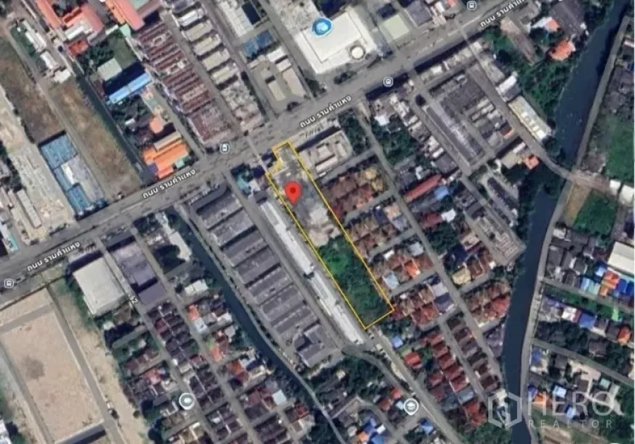 Land for sale in Bang Kapi, Bangkok - Entrance area of the cleared plot beside an industrial building and trees, flags visible on the roofline.