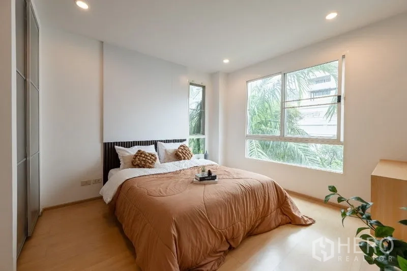 Condominium for sale in Watthana, Bangkok - Corner master bedroom with large windows, leafy views and warm-toned bedding.