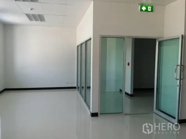 货栈 出租在Samut Prakan - 玻璃隔断的办公走廊，滑动门与醒目的紧急出口指示。