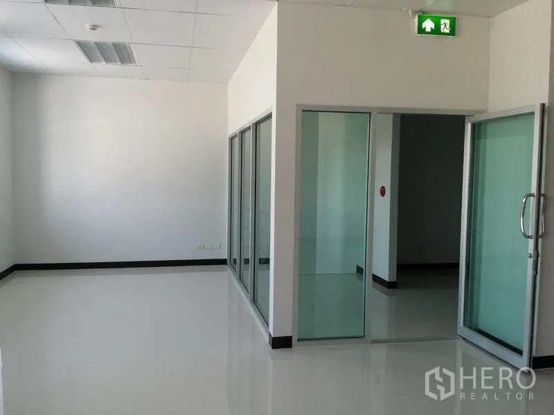 货栈 出租在Samut Prakan - 玻璃隔断的办公走廊，滑动门与醒目的紧急出口指示。