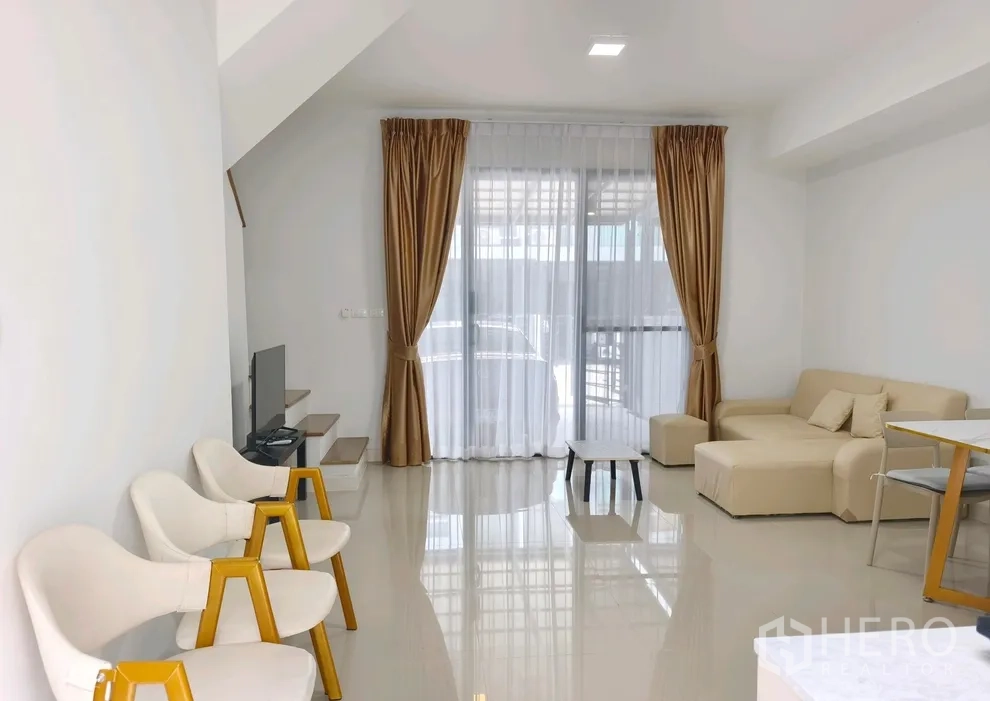Townhouse for rent in Bang Phli, Samut Prakan - Living room with L-shaped sofa, TV and full-height curtains opening to the carport.