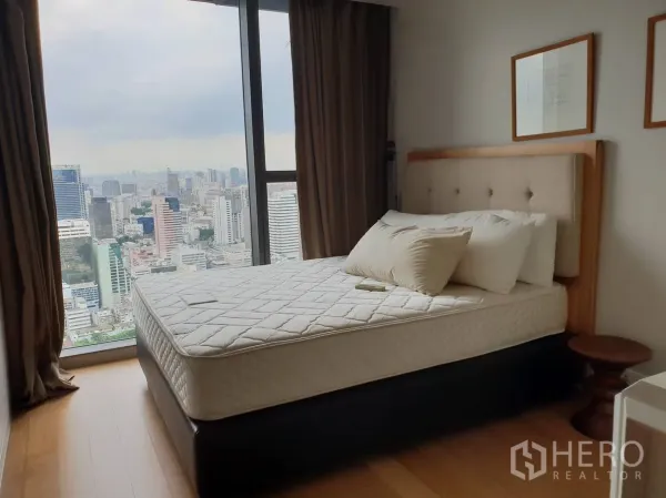 Condominium for rent in Sathon, Bangkok - Bedroom with floor-to-ceiling window offering panoramic Bangkok skyline views, wood floors and a padded-headboard bed.